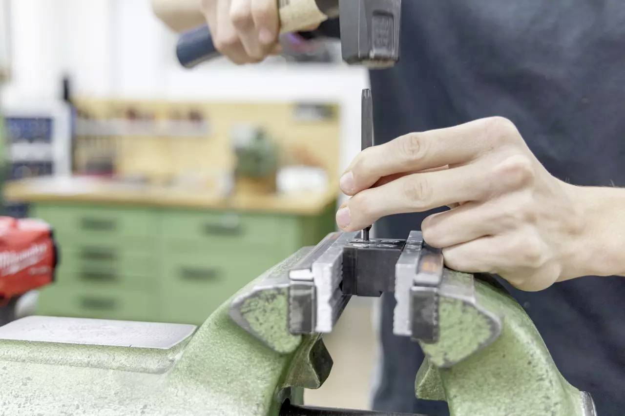Eine Person, die einen Hammer und einen Meißel an einem in einem Schraubstock befestigten Metallstück in einer Werkstattumgebung verwendet und sich auf detailliertes Handwerk konzentriert.