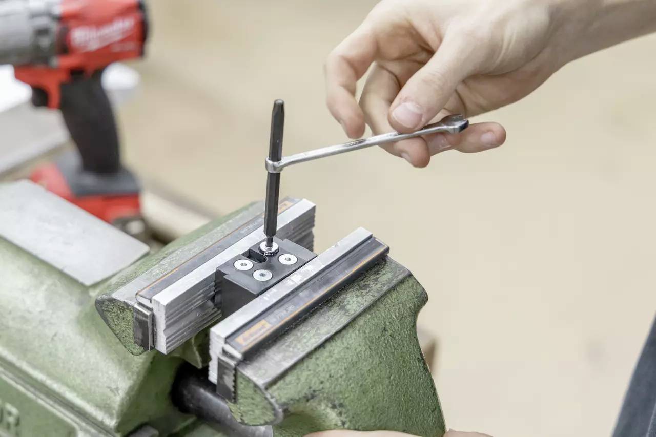 Eine Person benutzt einen Schraubenschlüssel, um eine Schraube an einer Komponente zu befestigen, die in einem Metallschraubstock eingespannt ist, wobei im Hintergrund ein Akkuschrauber zu sehen ist.
