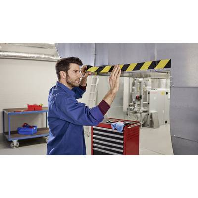 Ein Mann in Arbeitskleidung bringt ein Warnband an einer niedrigen Metallkante in einem industriellen Raum an. Im Hintergrund sind Werkzeuge zu sehen.