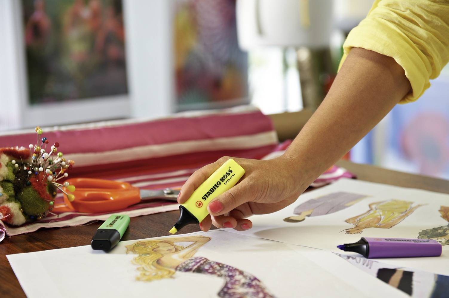 Eine Person markiert farbenfrohe Modezeichnungen auf einem Tisch mit Textmarkern. Daneben liegen bunte Stoffe und Scheren.