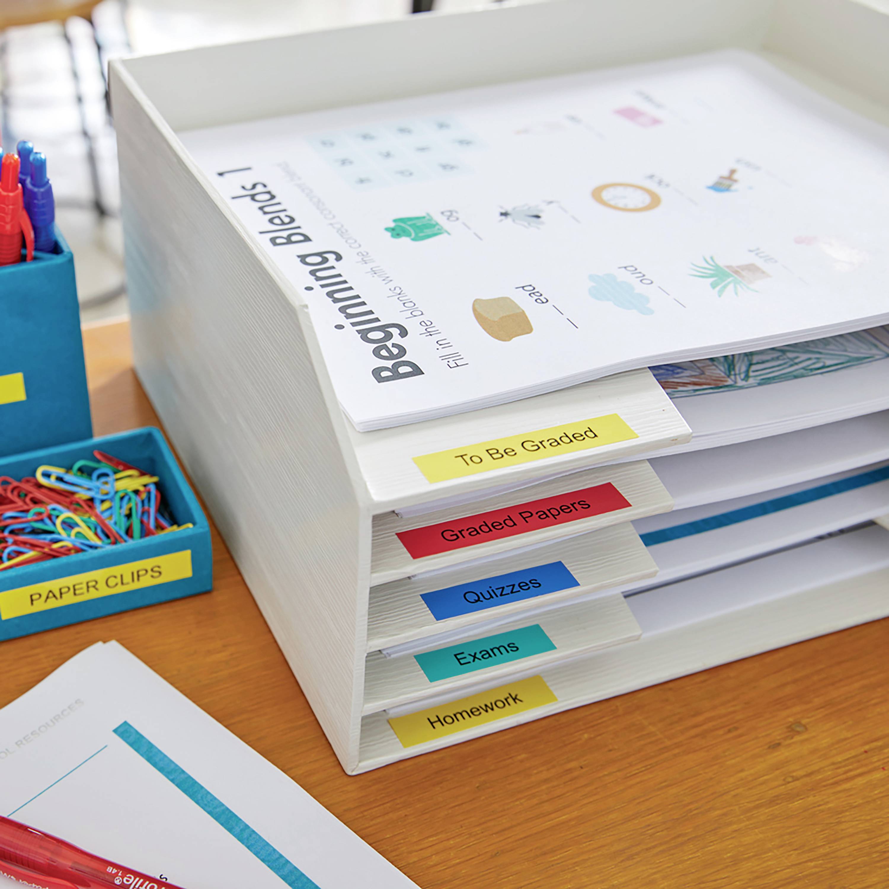 Ein Dokumentenablagefach mit mehreren Fächern, beschriftet mit 'To Be Graded', 'Graded Papers', 'Tests', 'Quizzes', 'Exams', 'Homework'. Ein Arbeitsblatt mit 'Beginning Blends' liegt darauf.