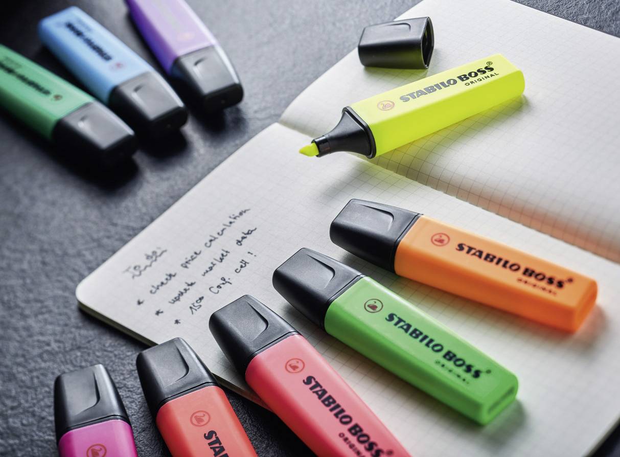 Ein Notizbuch auf einem Tisch, umgeben von bunten Textmarkern in Grün, Blau, Lila, Gelb, Orange und Pink.