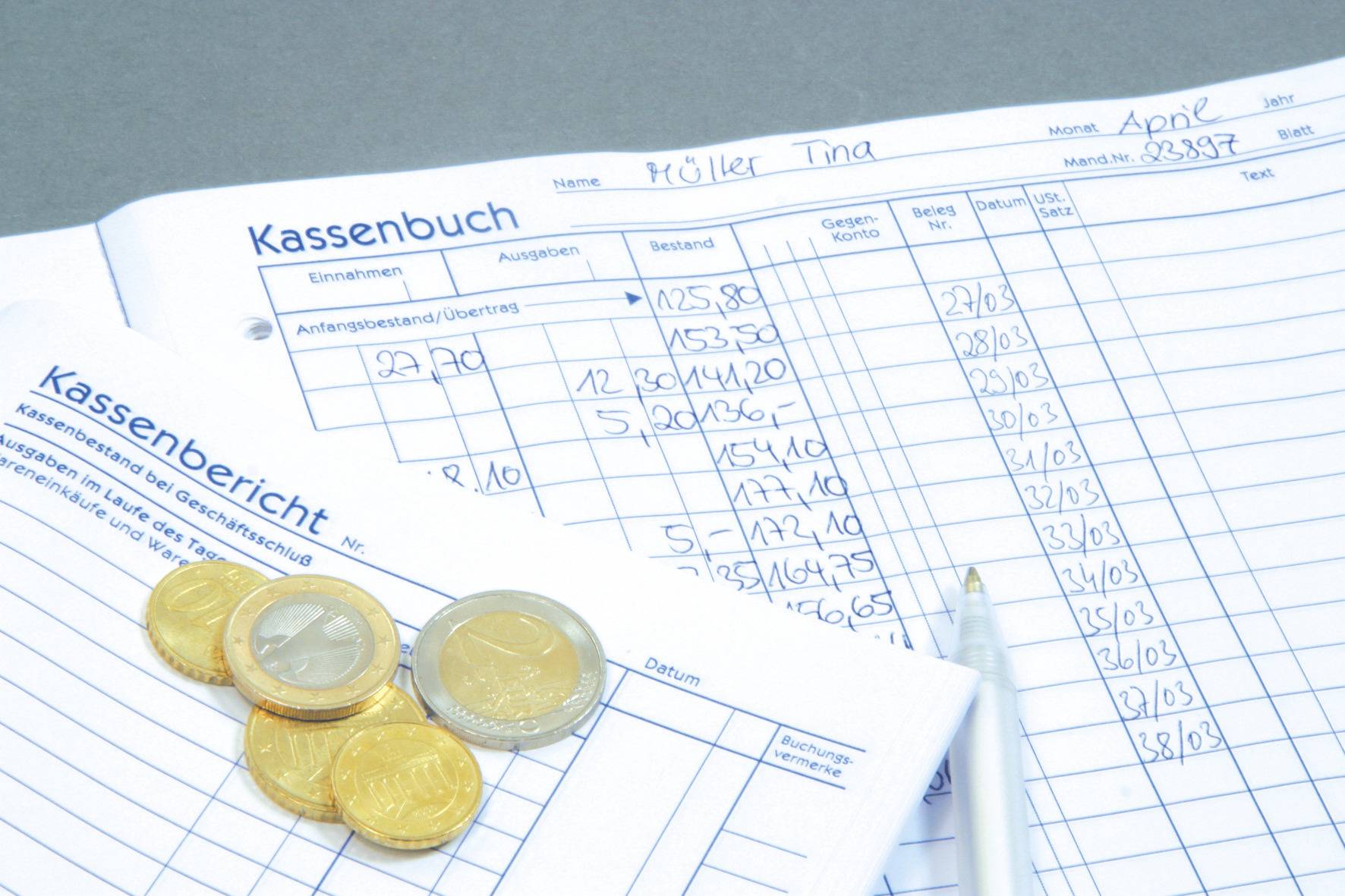 Ein Kassenbuch mit handgeschriebenen Zahlen und einem Kassenbericht liegt auf einem Tisch. Daneben befinden sich Euro-Münzen und ein Stift.