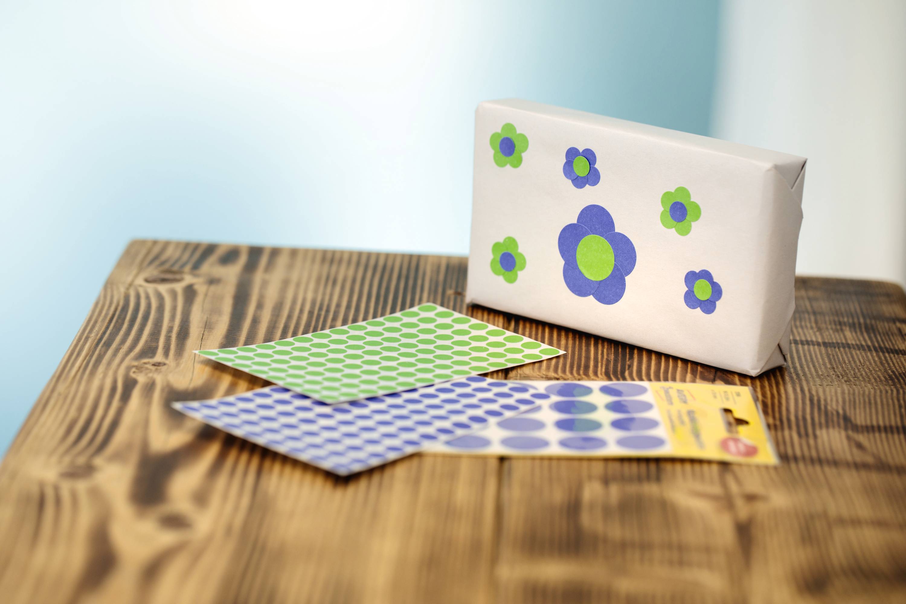 Ein verpacktes Geschenk mit Blumenmotiven steht auf einem Holztisch, daneben liegen grüne und blaue Punkteaufkleber in verschiedenen Mustern.