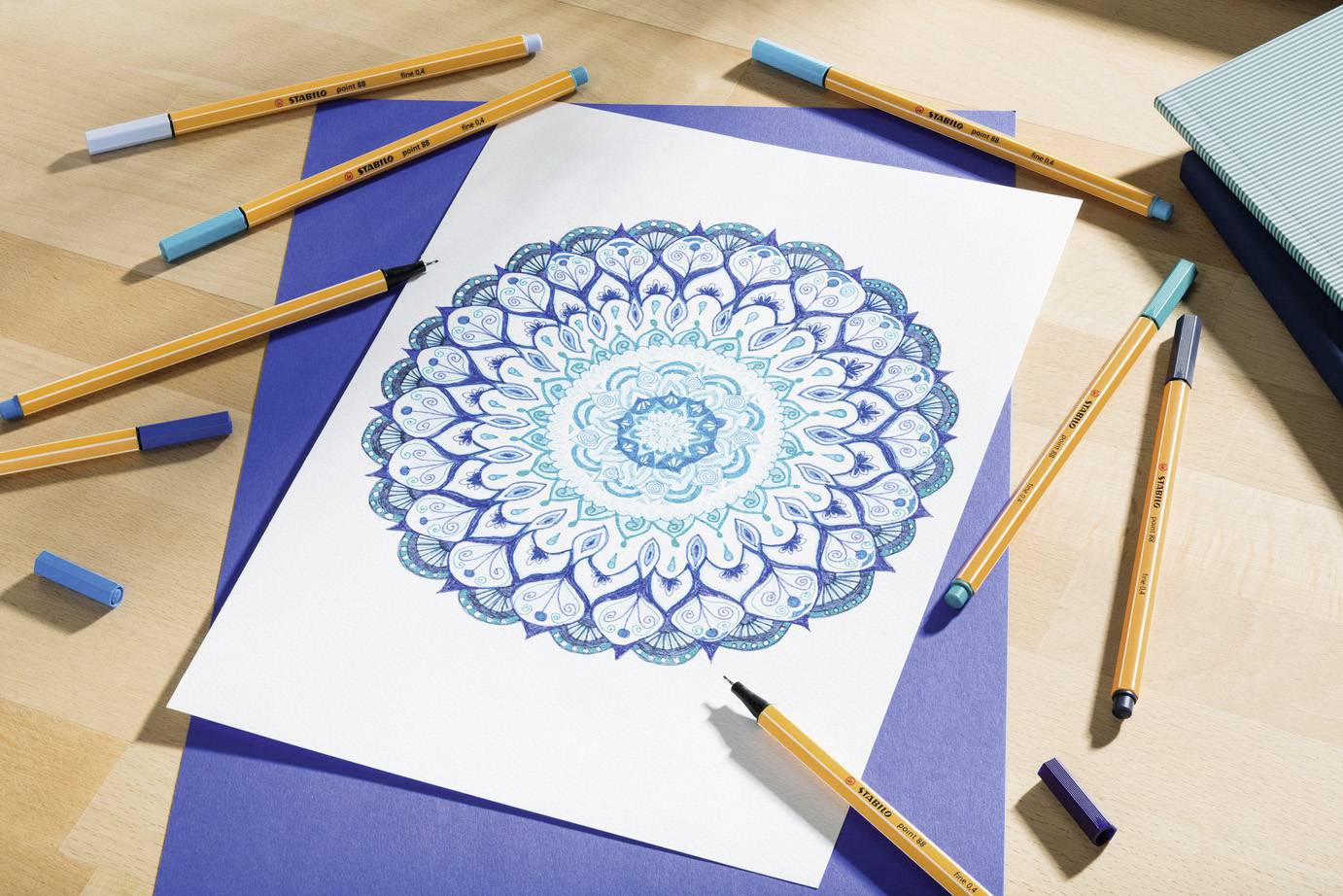Ein Mandala in Blau- und Türkistönen liegt auf einem hellen Tisch, umgeben von Stiften. Es zeigt ein symmetrisches, kreisförmiges Muster.