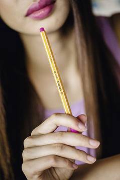 Eine Person hält einen gelb-pinken Stift zwischen den Fingern. Der Fokus liegt auf dem Stift, der leicht nach oben gehalten wird.