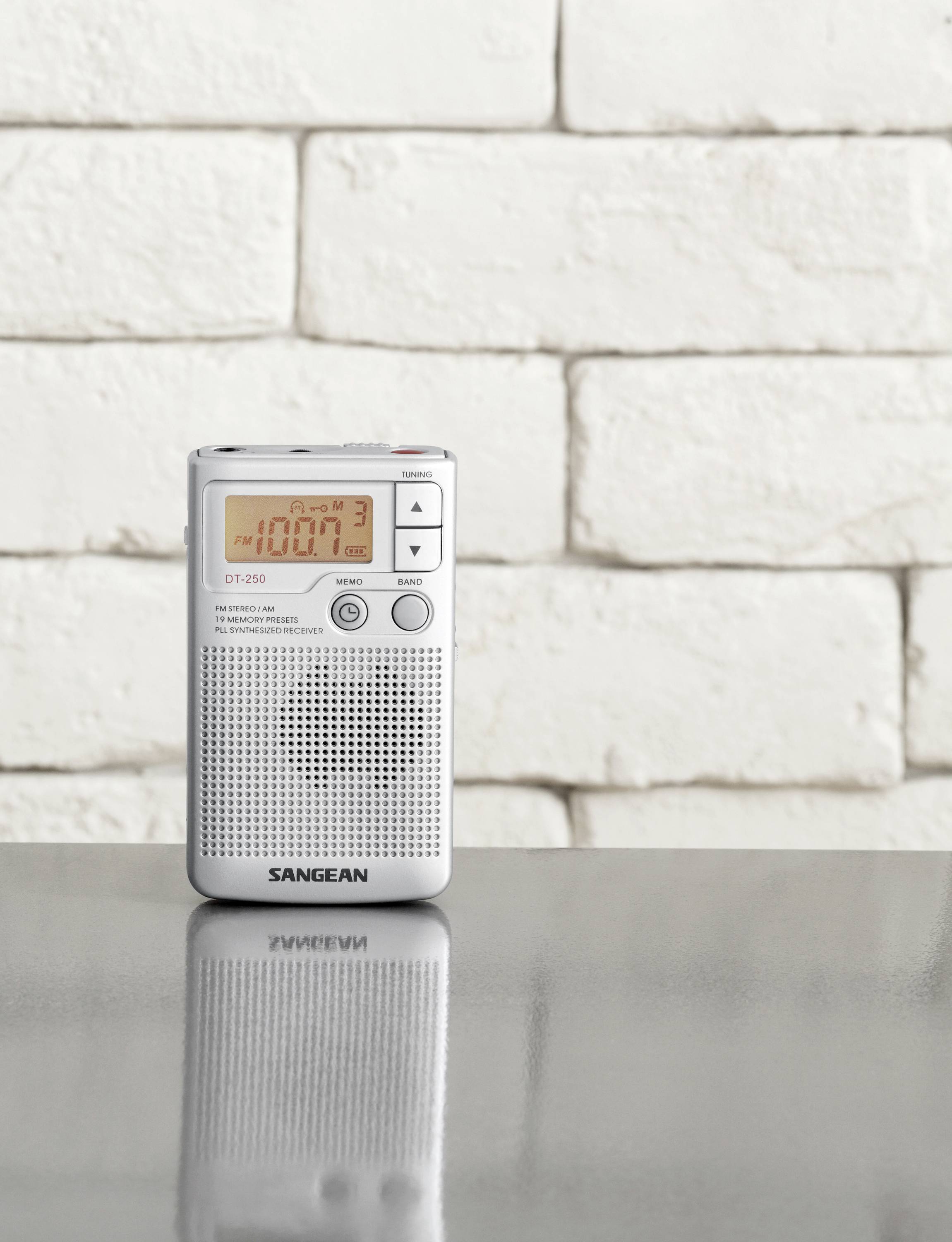 Ein kleines tragbares Radio steht auf einem Tisch vor einer weißen Ziegelwand. Es zeigt 100.7 auf dem Display an.