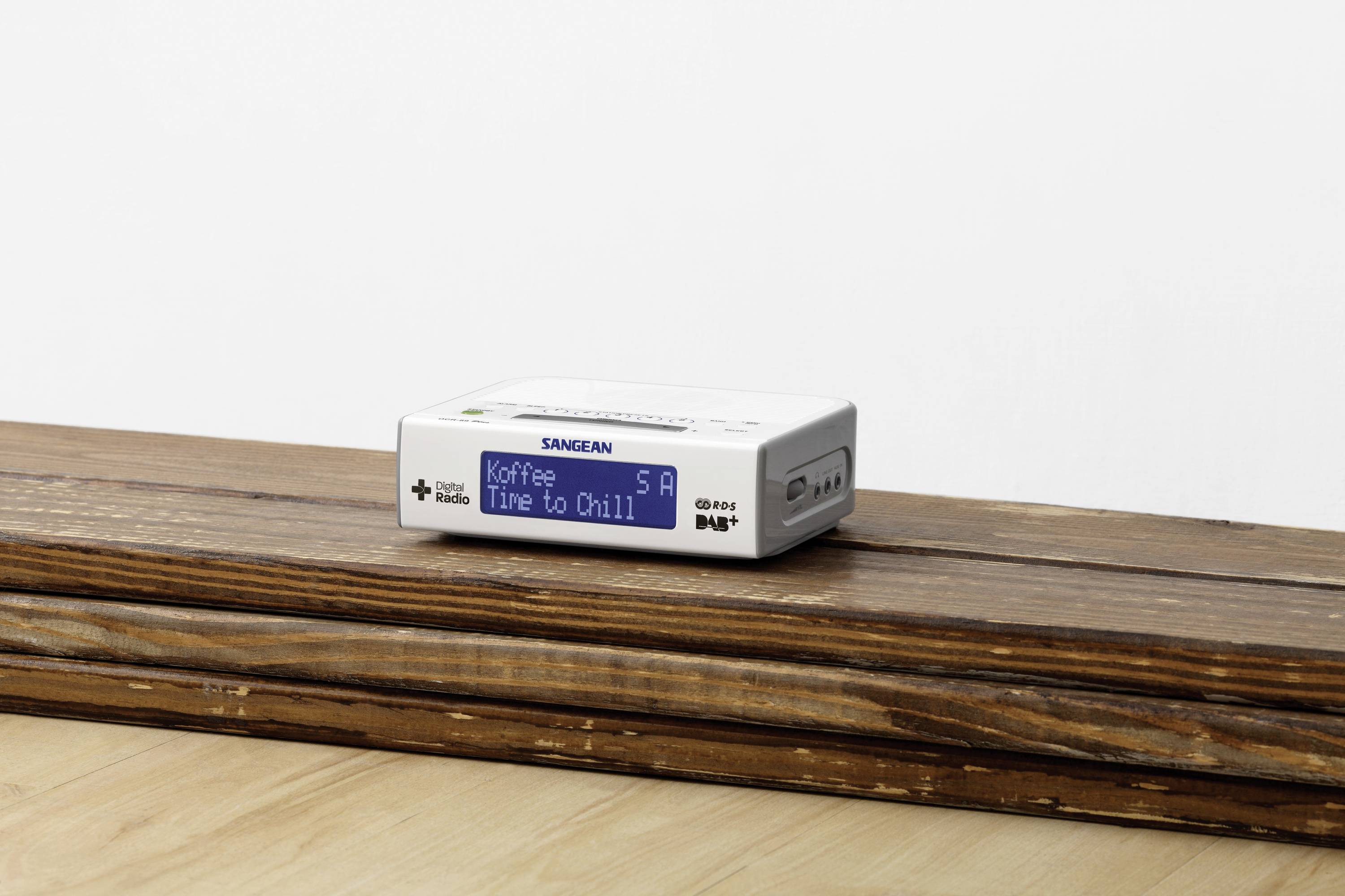 Ein kleines, weißes tragbares Radio auf einem Holztisch zeigt auf dem Display 'Koffee Time to Chill'.
