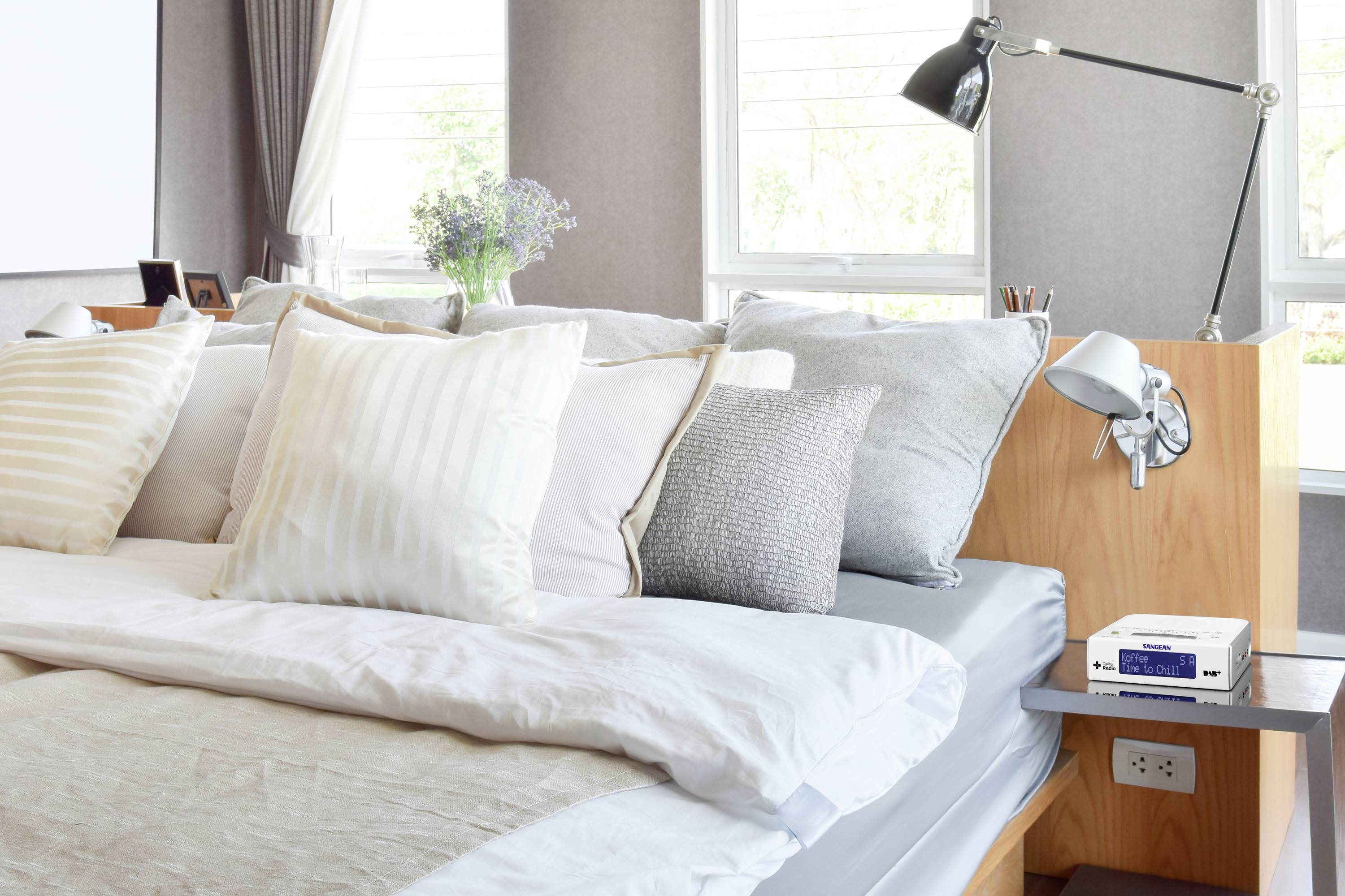 Helles Schlafzimmer mit einem großen, gemachten Bett, dekoriert mit verschiedenen Kissen und einer Vase mit Blumen auf dem Kopfteil.