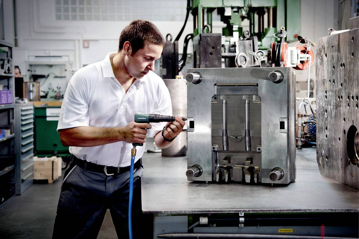 Ein Mann in einem weißen Hemd arbeitet in einer Werkstatt mit einem Druckluftschrauber an einer großen Metallmaschine.