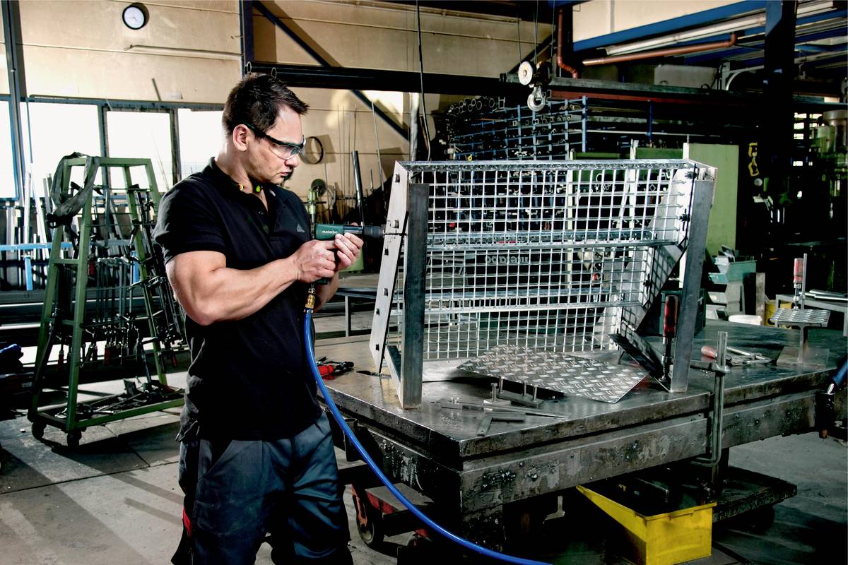 Ein Mann reinigt in einer Werkstatt mit einem Druckluftgerät ein Metallgitter. Im Hintergrund sind Maschinen und Werkzeuge sichtbar.
