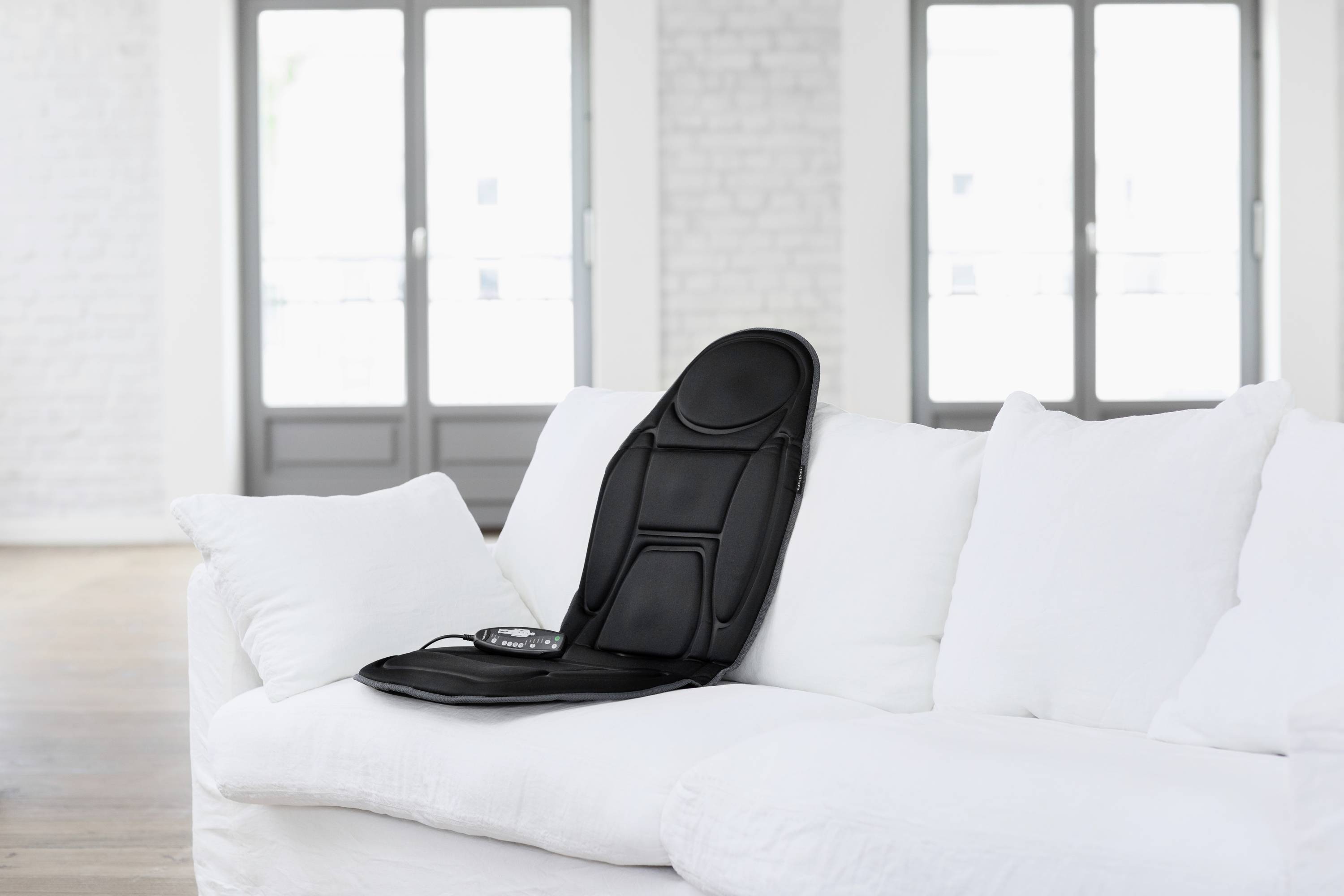 Eine schwarze Massagematte liegt auf einem weißen Sofa in einem hellen Raum mit großen Fenstern im Hintergrund.