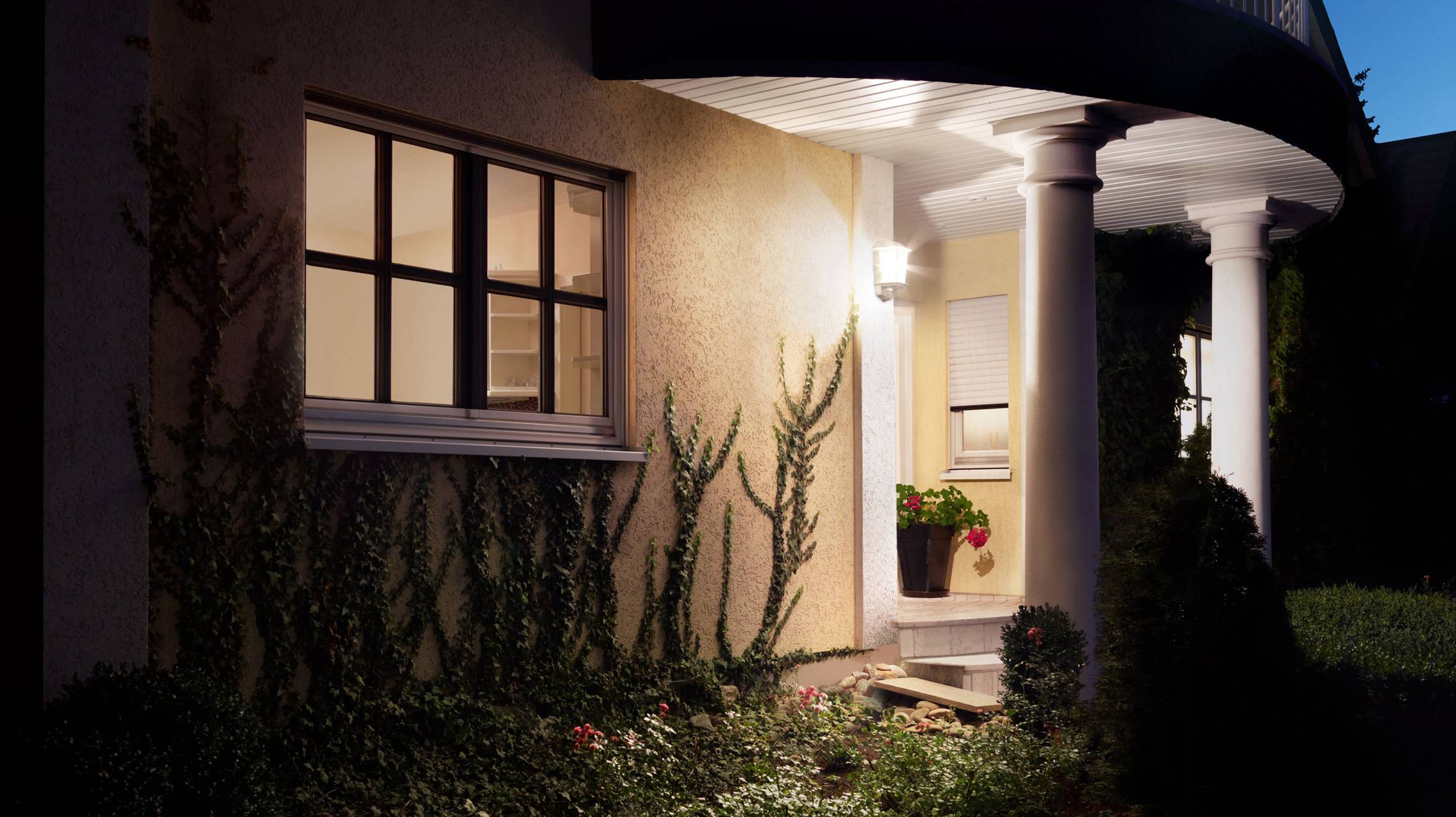 Ein beleuchteter Hauseingang bei Nacht mit zwei Säulen, beleuchtetes Fenster und Pflanzen im Vordergrund.