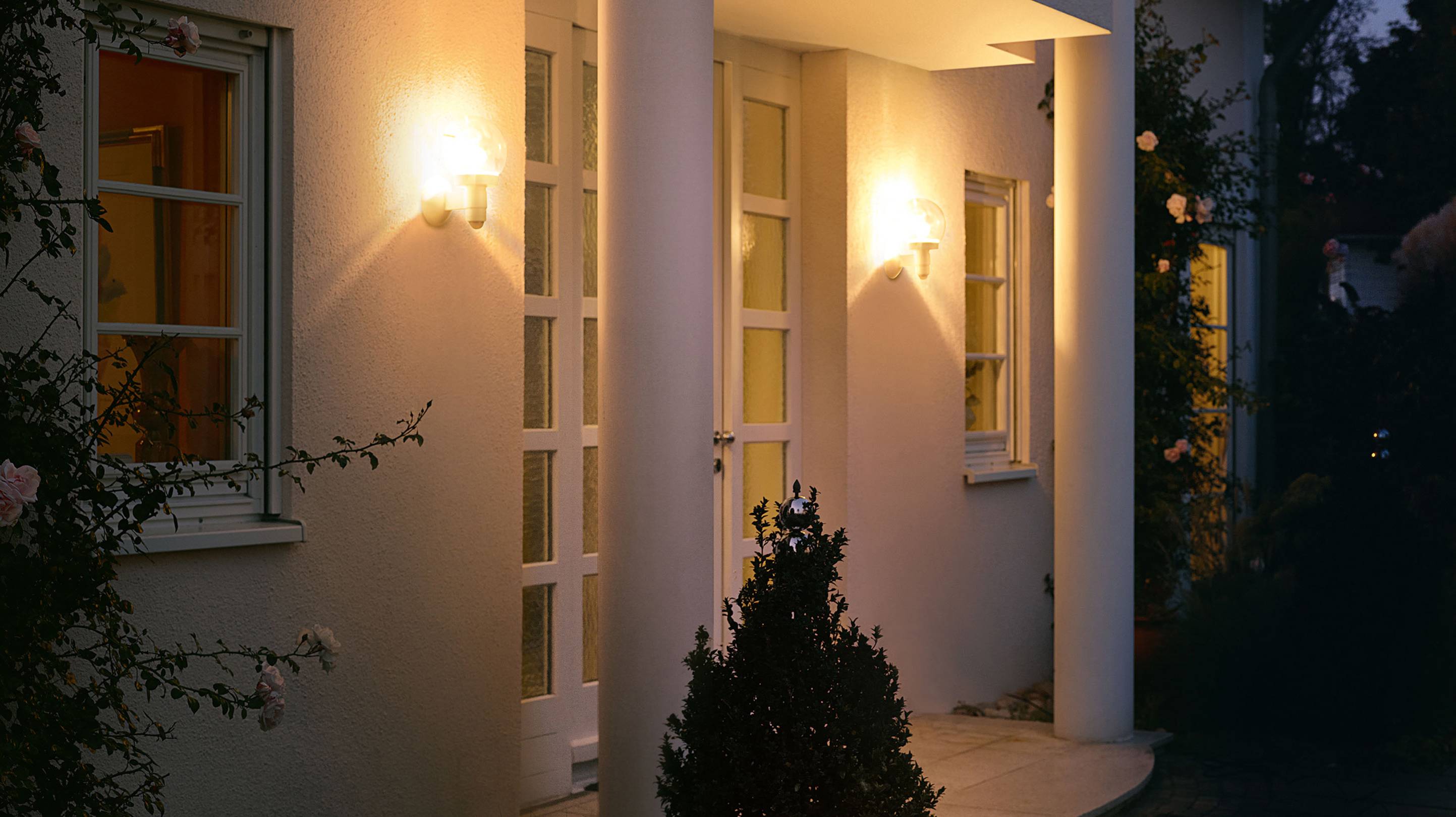 Eingangsbereich eines Hauses bei Dämmerung, beleuchtet von warmen Wandlampen. Weiße Säulen, Fenster mit klassischen Holzrahmen.