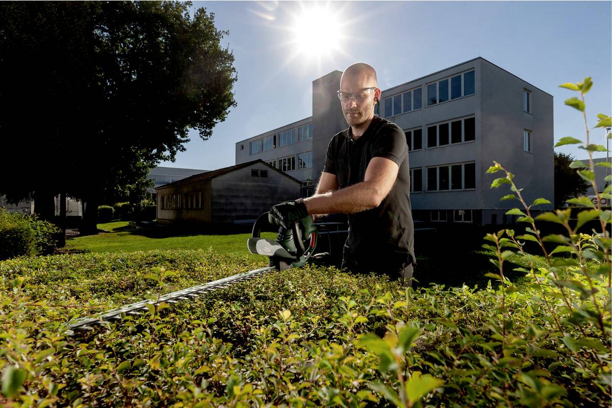 Ein Mann mit Schutzbrille schneidet eine Hecke mit einer Heckenschere vor einem modernen zweistöckigen Gebäude bei Sonnenschein.