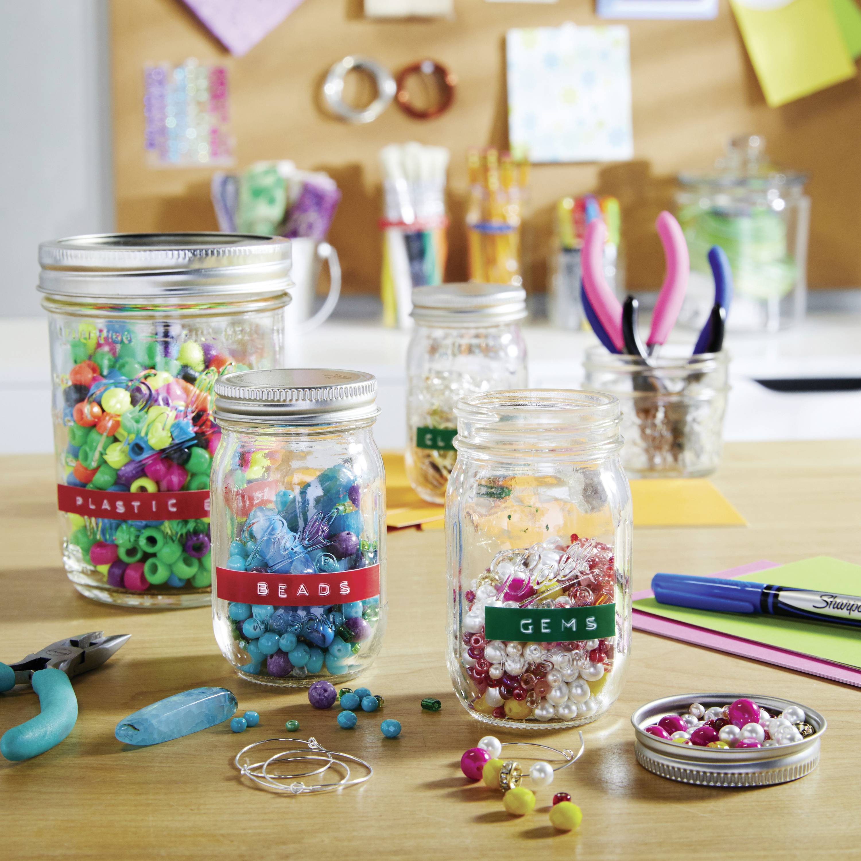 In einem Bastelraum stehen mehrere Glasbehälter mit bunten Perlen und Edelsteinen auf einem Holztisch, umgeben von Werkzeugen und Materialien.