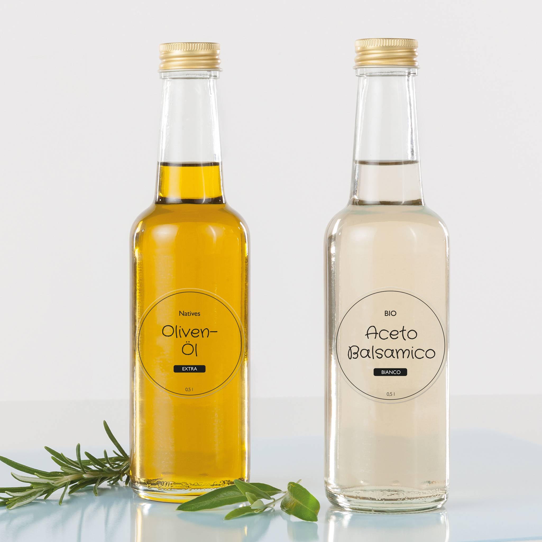 Zwei Glasflaschen auf einem Tisch mit Olivenzweigen, links Olivenöl mit goldener Flüssigkeit, rechts weißer Balsamessig.