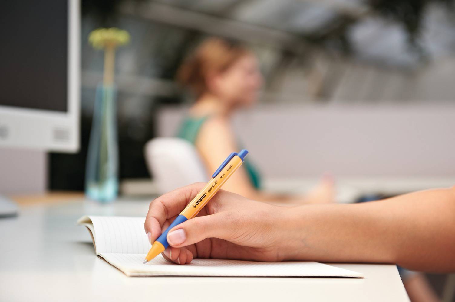 Eine Hand notiert in einem Notizbuch mit einem gelben Stift, im Hintergrund eine Person unscharf.