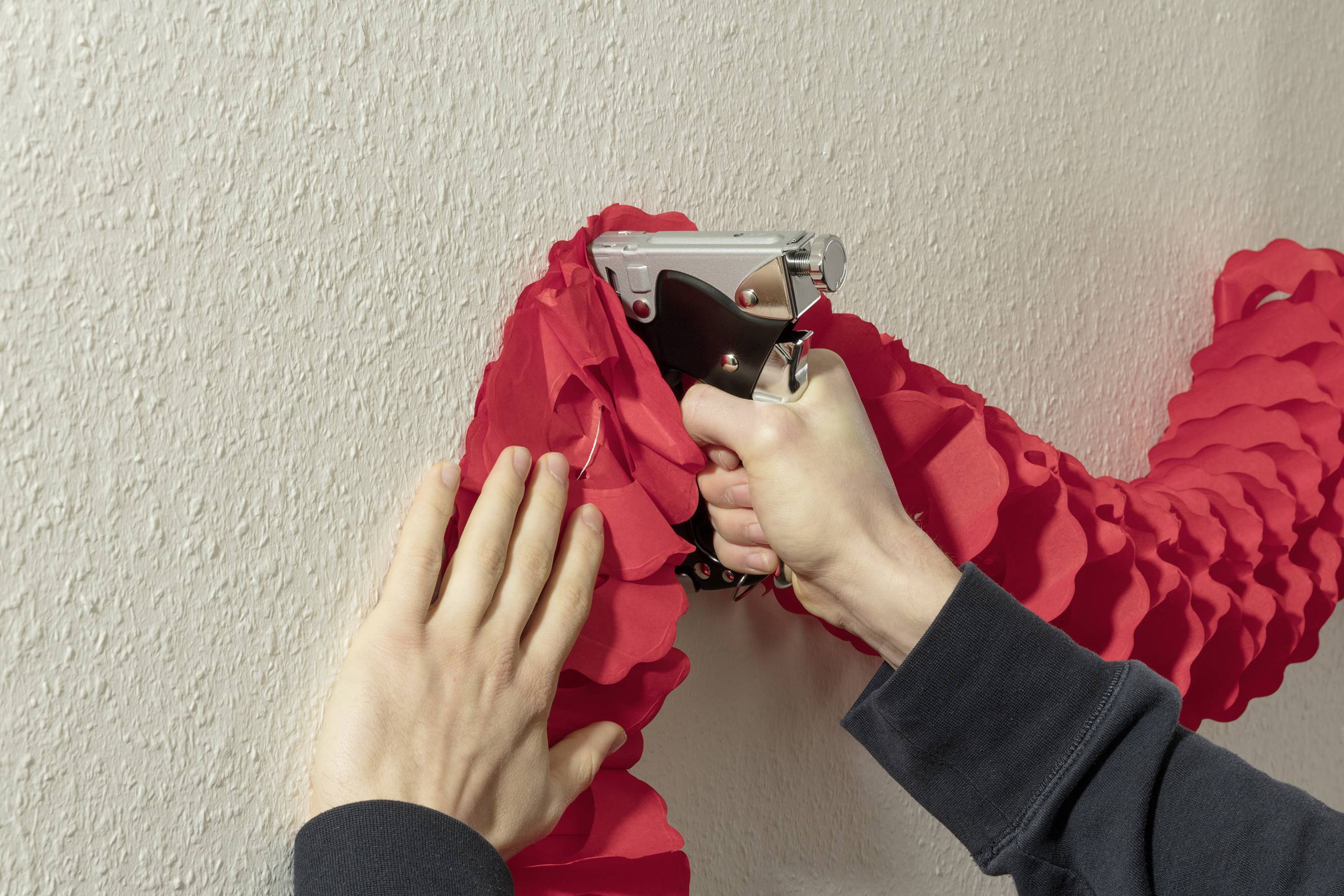 Eine Person befestigt eine rote Girlande mit einem Tacker an einer Wand. Hände bearbeiten das Material, um es sicher zu fixieren.