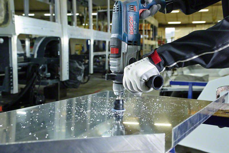 Eine Person bohrt mit einer elektrischen Bohrmaschine in ein Metallstück. Späne fliegen, und im Hintergrund sieht man eine Werkstattumgebung.