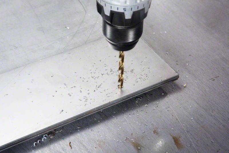 Ein Bohrer durchbohrt ein Stück Metall, wobei Späne auf der Oberfläche verteilt sind. Die Szene zeigt einen Arbeitsvorgang in der Metallbearbeitung.