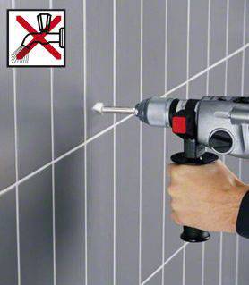 Eine Hand hält eine Bohrmaschine, die in eine graue Fliese bohrt. Oben links befindet sich ein Symbol, das das Nassbohren verbietet.