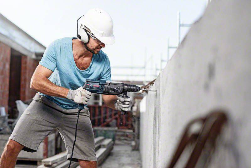 Ein Bauarbeiter mit Helm und Gehörschutz bohrt mit einer Bohrmaschine in eine Betonwand auf einer Baustelle.