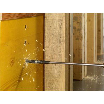 'Ein Bohrer durchdringt eine Holzwand, umrahmt von anderen Holzbalken. Sägespäne fliegen durch die Luft.'
