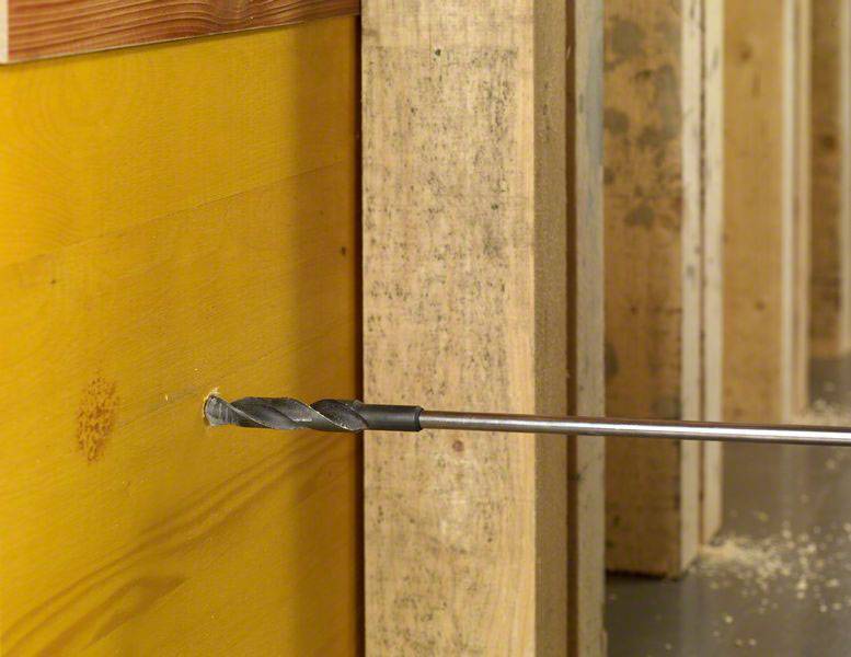 'Ein Bohrer durchdringt eine hölzerne Wand, wobei Holzspäne auf dem Boden sichtbar sind. Der Bohrer ist mit einem langen Metallstift verbunden.'