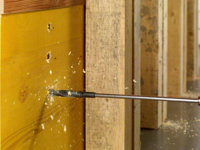 Eine Bohrmaschine durchdringt Holz, wobei Späne herausfliegen. Im Hintergrund sind mehrere Holzplatten sichtbar.