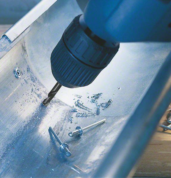 Eine Bohrmaschine durchbohrt ein Metallblech. Rund um das Bohrloch sind Metallspäne sichtbar. Daneben liegen Niete.