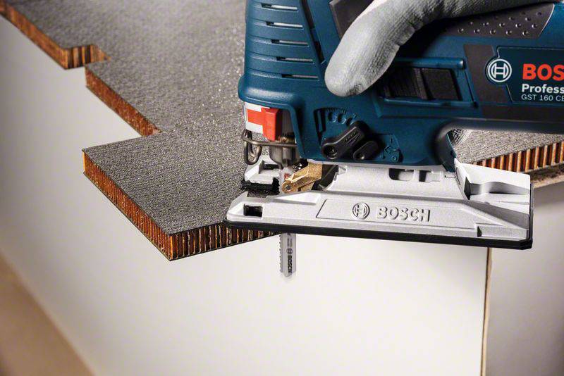 Eine Person sägt mit einer blauen Stichsäge von Bosch durch eine mehrschichtige Holzplatte. Die Säge gleitet präzise entlang der Kante.