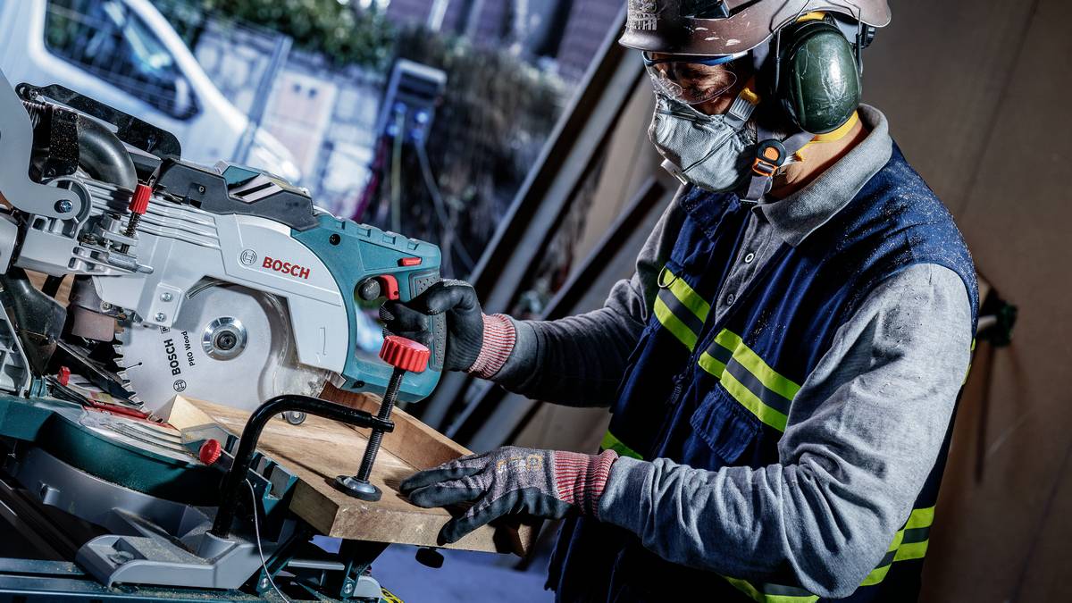 Ein Schreiner mit Schutzausrüstung verwendet eine Bosch-Kreissäge, um in einer Werkstattumgebung ein Stück Holz zu schneiden.