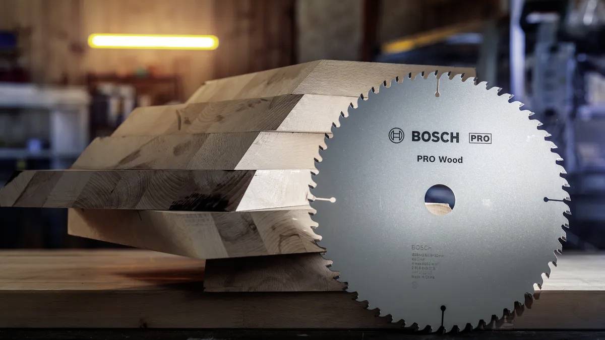 Eine Kreissägeblatt mit der Aufschrift „BOSCH PRO Wood", das vor gestapelten Holzplanken in einer Werkstattumgebung präsentiert wird.