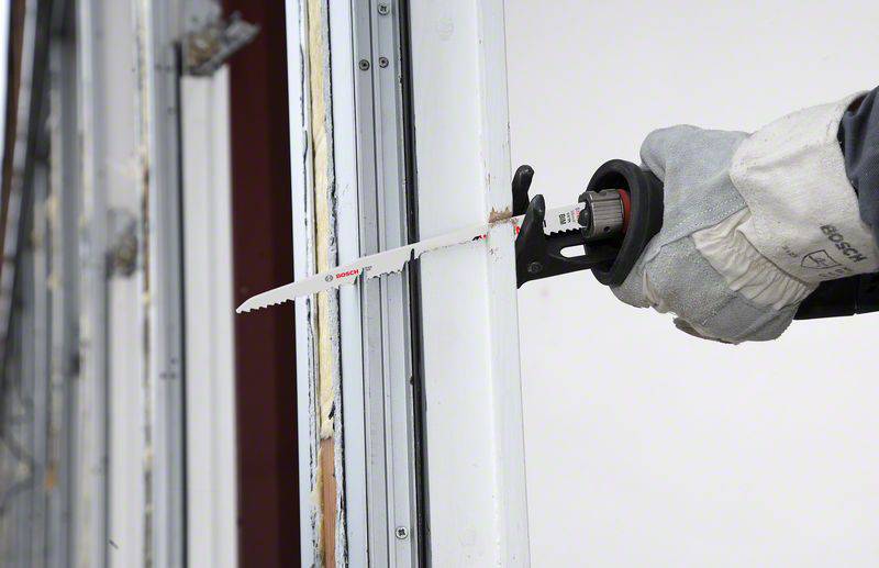 Hand mit Arbeitshandschuh hält elektrische Säge und schneidet Metallrahmen. Sägeblatt ist lang und scharf, Einsatz auf Baustelle.