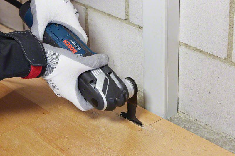 Eine Person schneidet mit einem oszillierenden Werkzeug eine Holzplatte an der Kante einer Wand. Sie trägt Handschuhe zum Schutz.