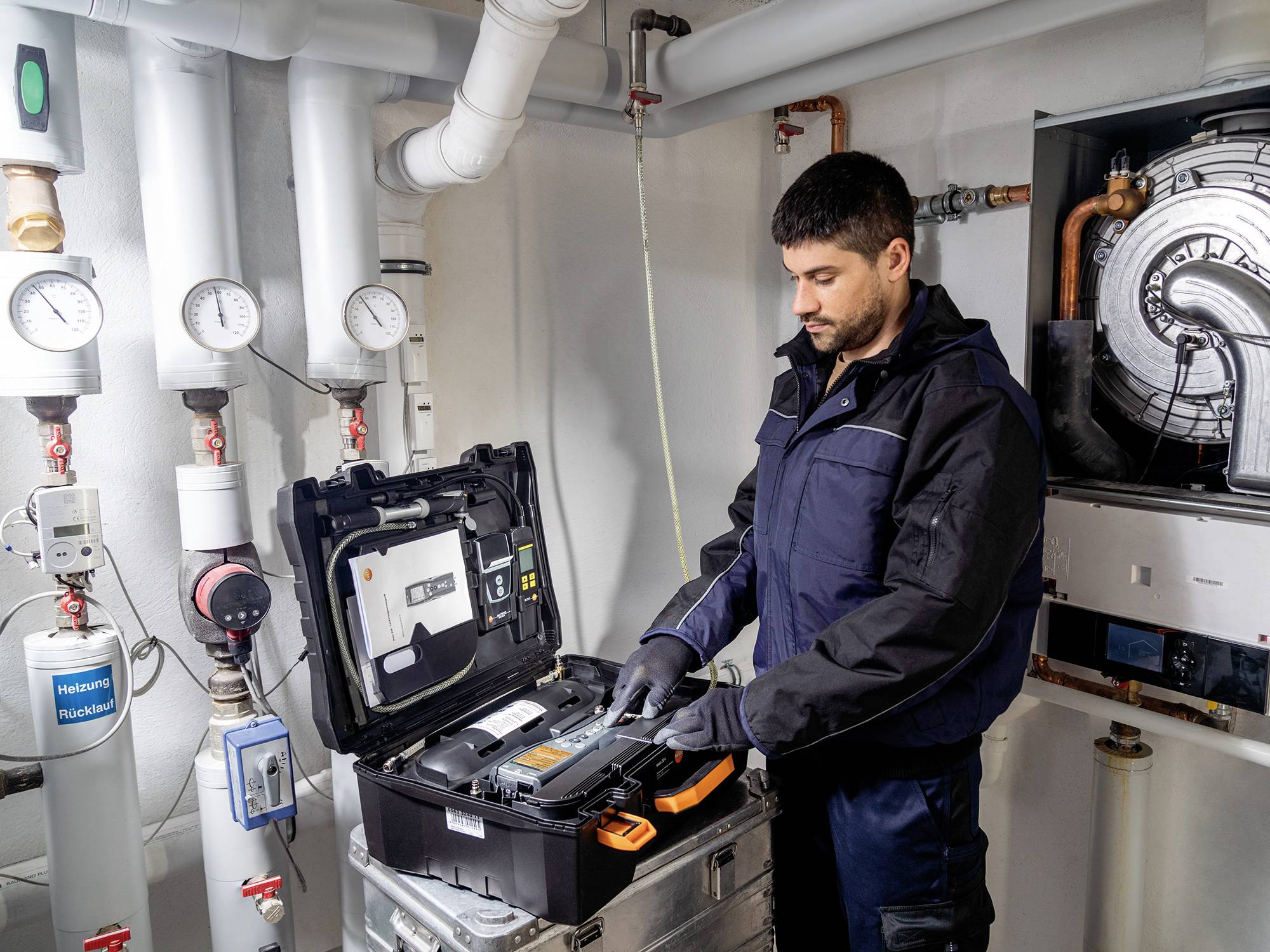 Ein Techniker arbeitet in einem Heizungsraum an einem geöffneten Koffer mit Instrumenten. Im Hintergrund sind Rohre und Messgeräte sichtbar.
