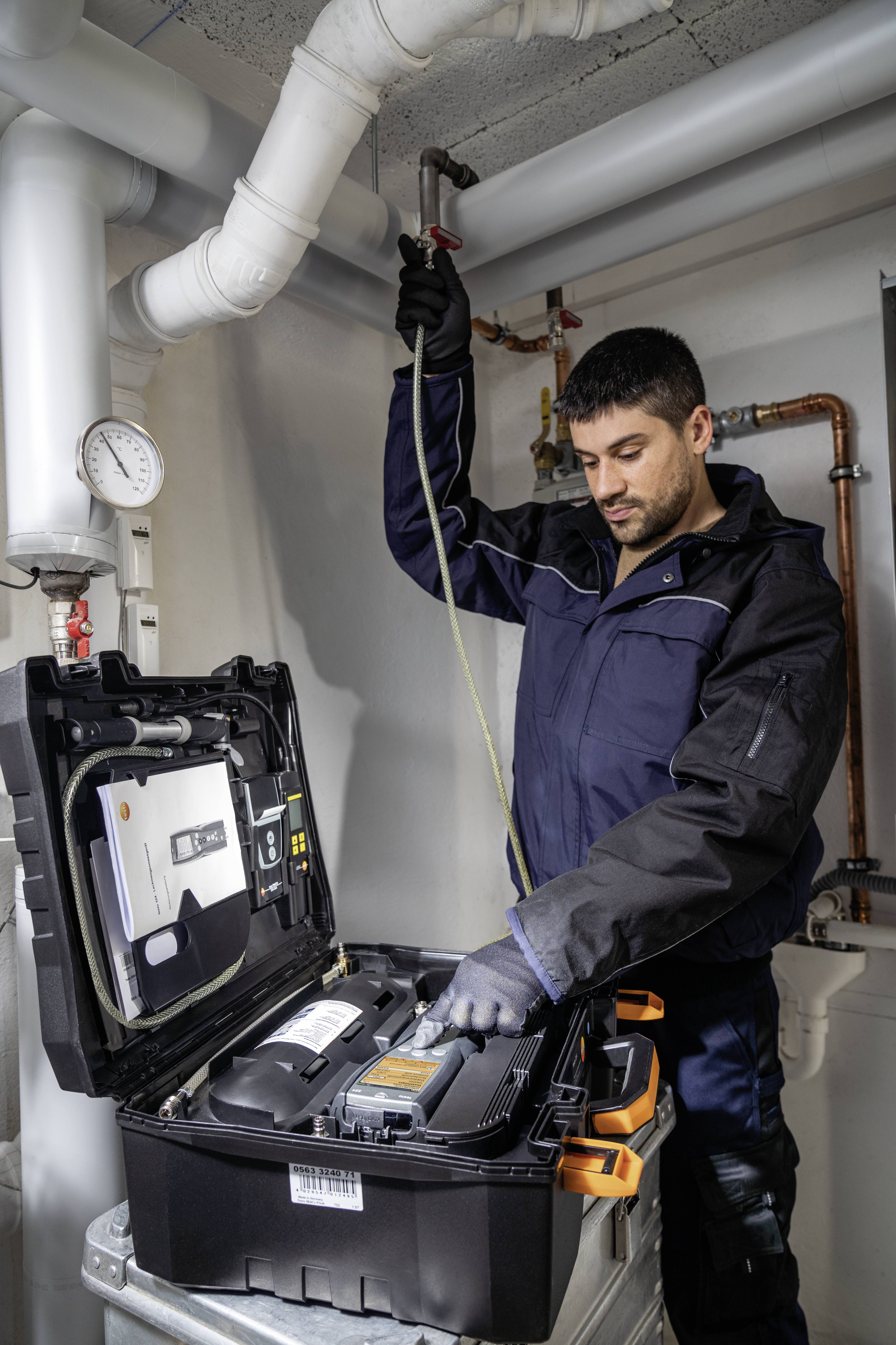 Ein Techniker in Arbeitskleidung überprüft ein Rohrsystem mit einem Messgerät. Er steht neben einem offenen Werkzeugkoffer in einem Heizungsraum.