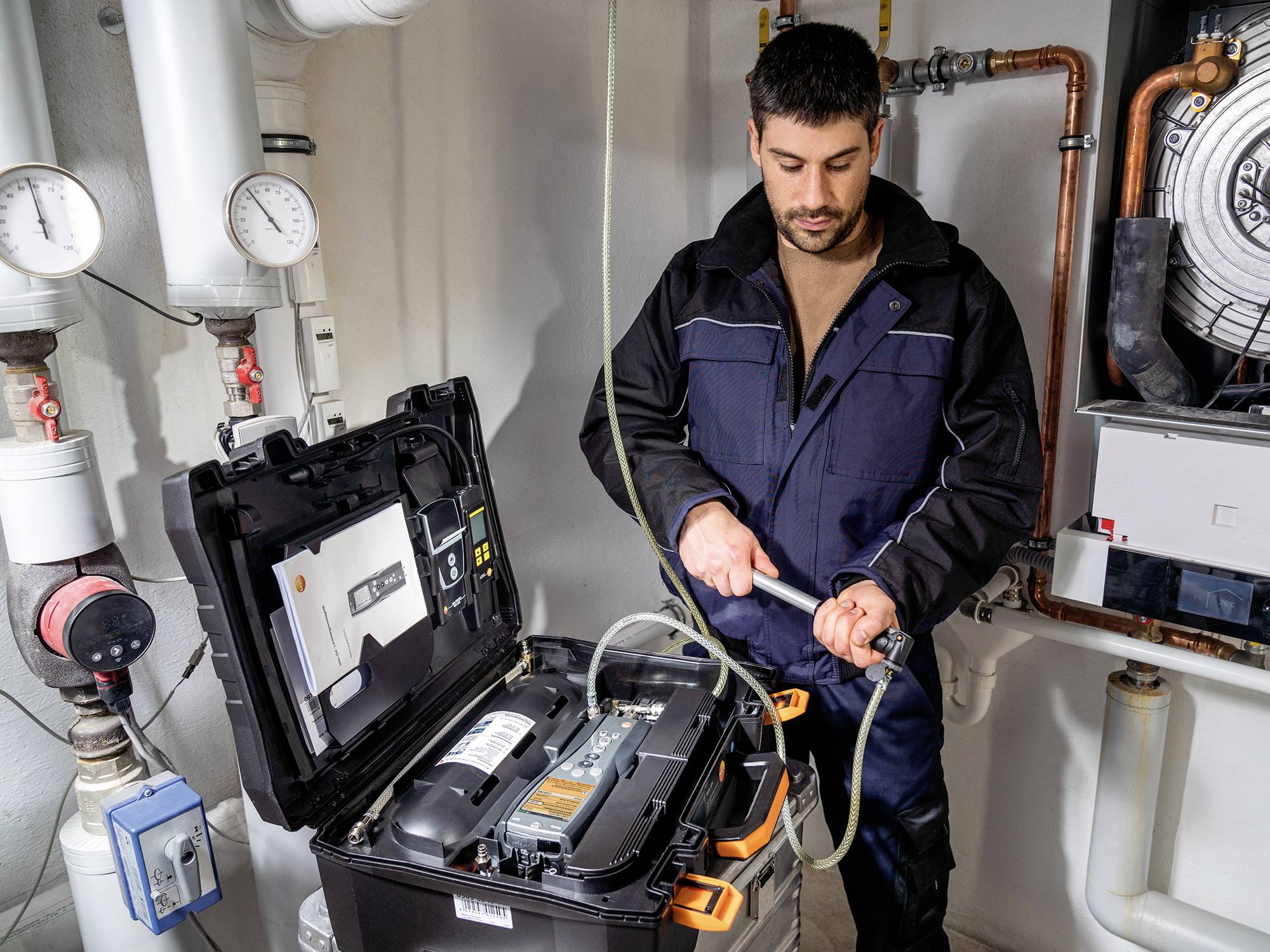 Ein Mann in Arbeitskleidung repariert ein technisches Gerät in einem Kellerraum. Er hält ein Werkzeug und ist konzentriert bei der Arbeit.