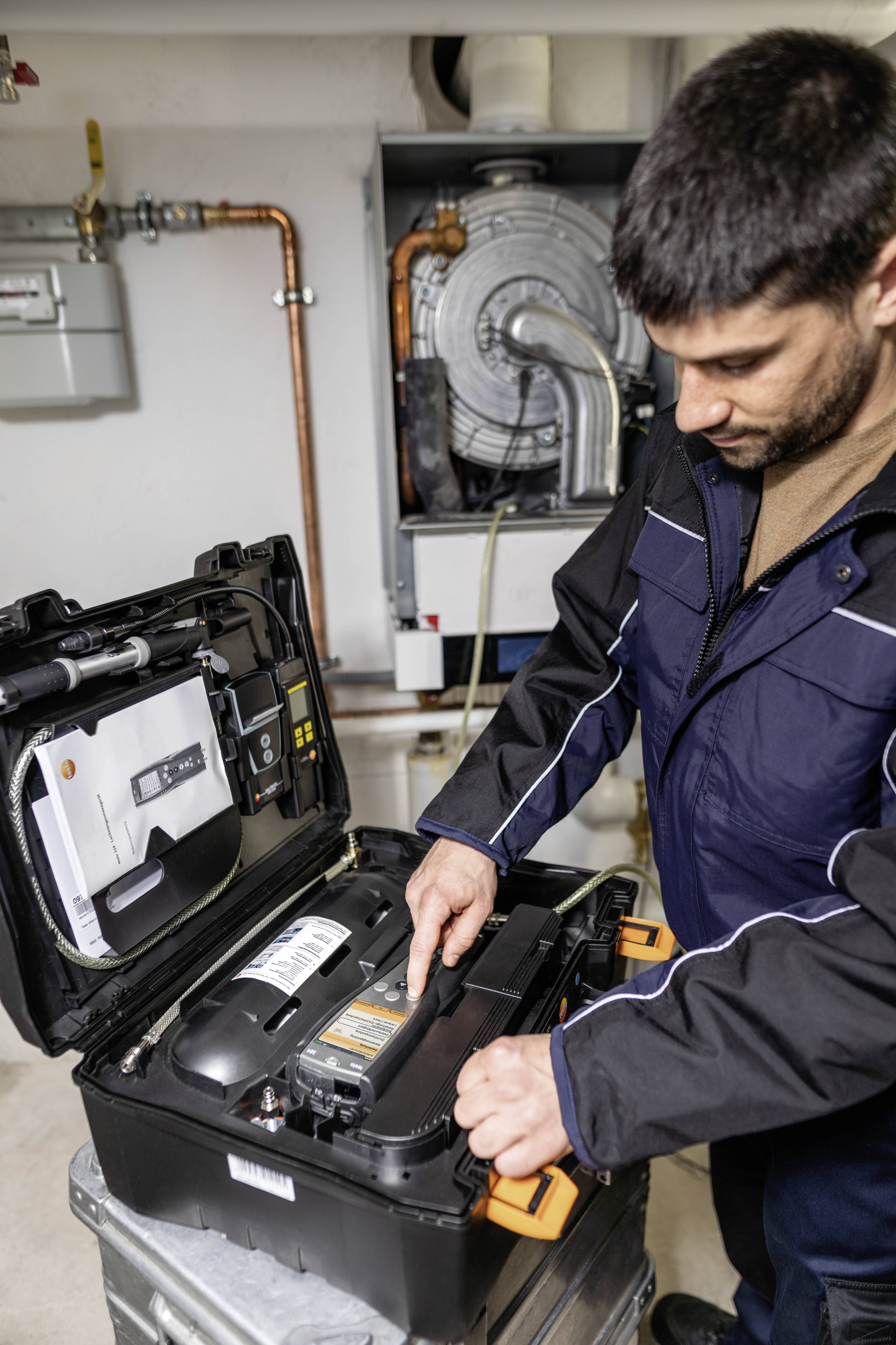 Ein Mann im Overall überprüft Geräte in einem Kofferraum in einem Technikraum. Im Hintergrund sind Rohre und eine Maschine sichtbar.