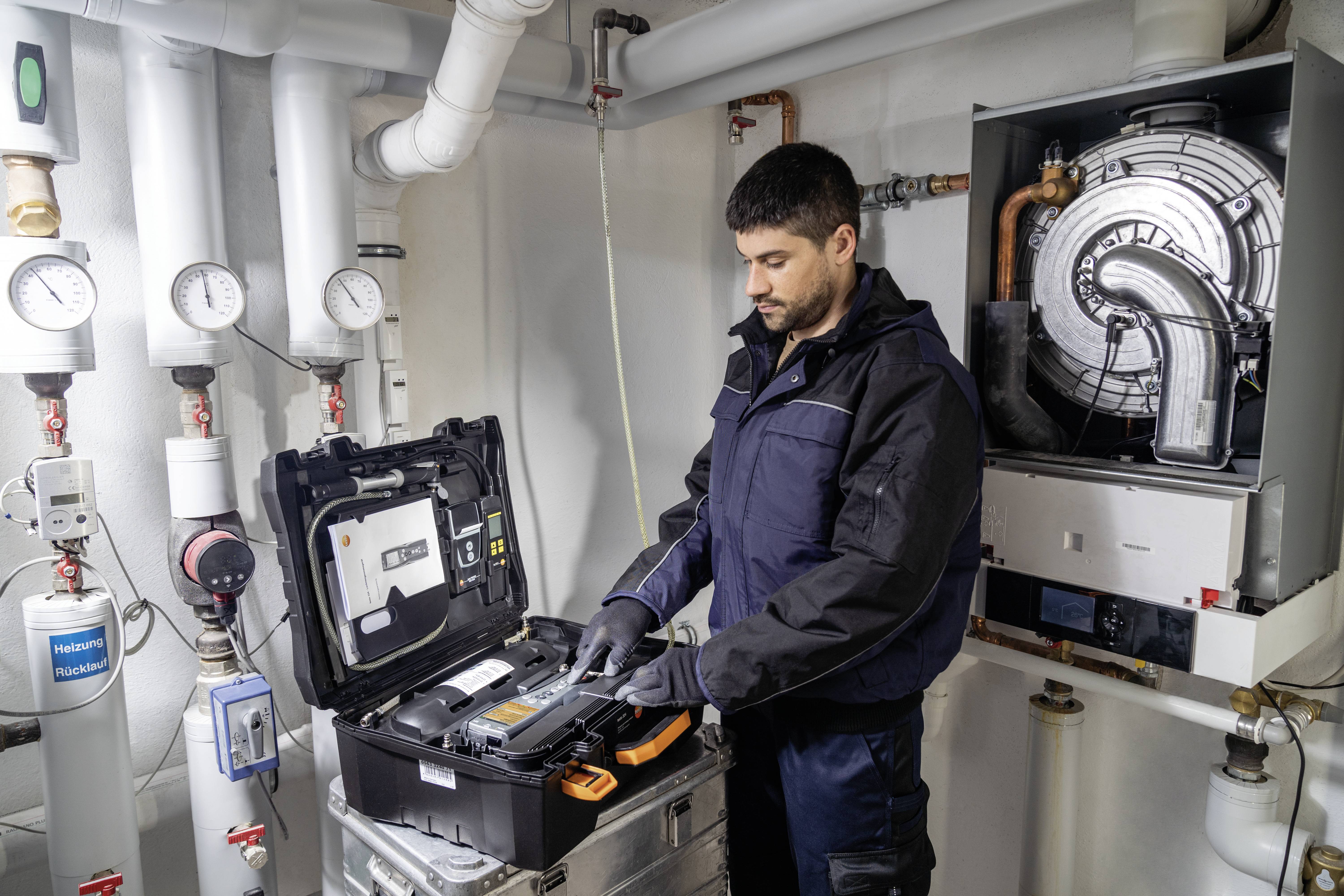Ein Techniker wartet ein Heizsystem in einem technischen Raum, umgeben von Rohren und Manometern. Er arbeitet mit einem Werkzeugkasten.