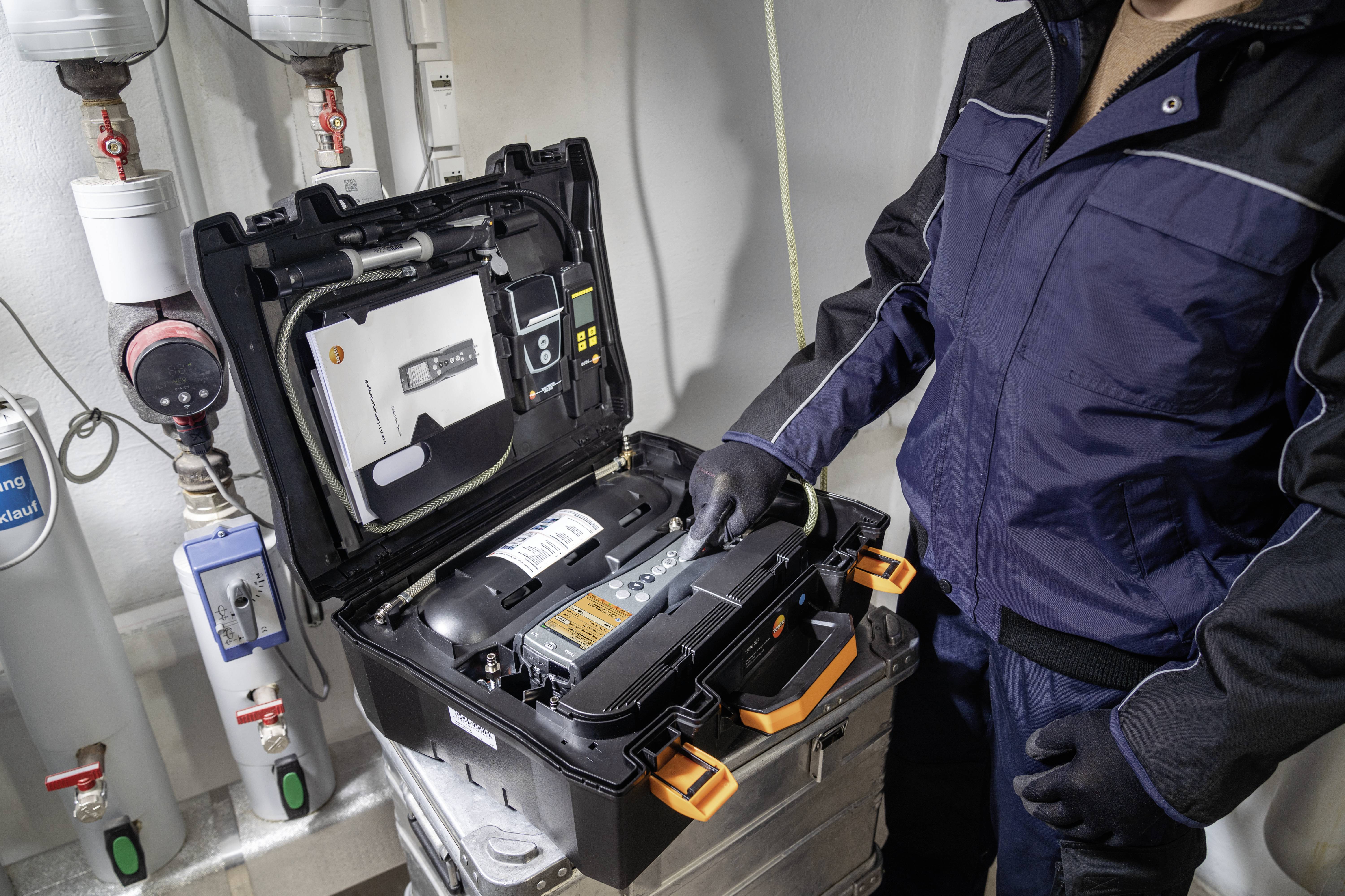 Person in Arbeitskleidung überprüft ein offenes technisches Gerät in einem Gebäude. Es ist offenbar Teil einer Wartungs- oder Reparaturarbeit.