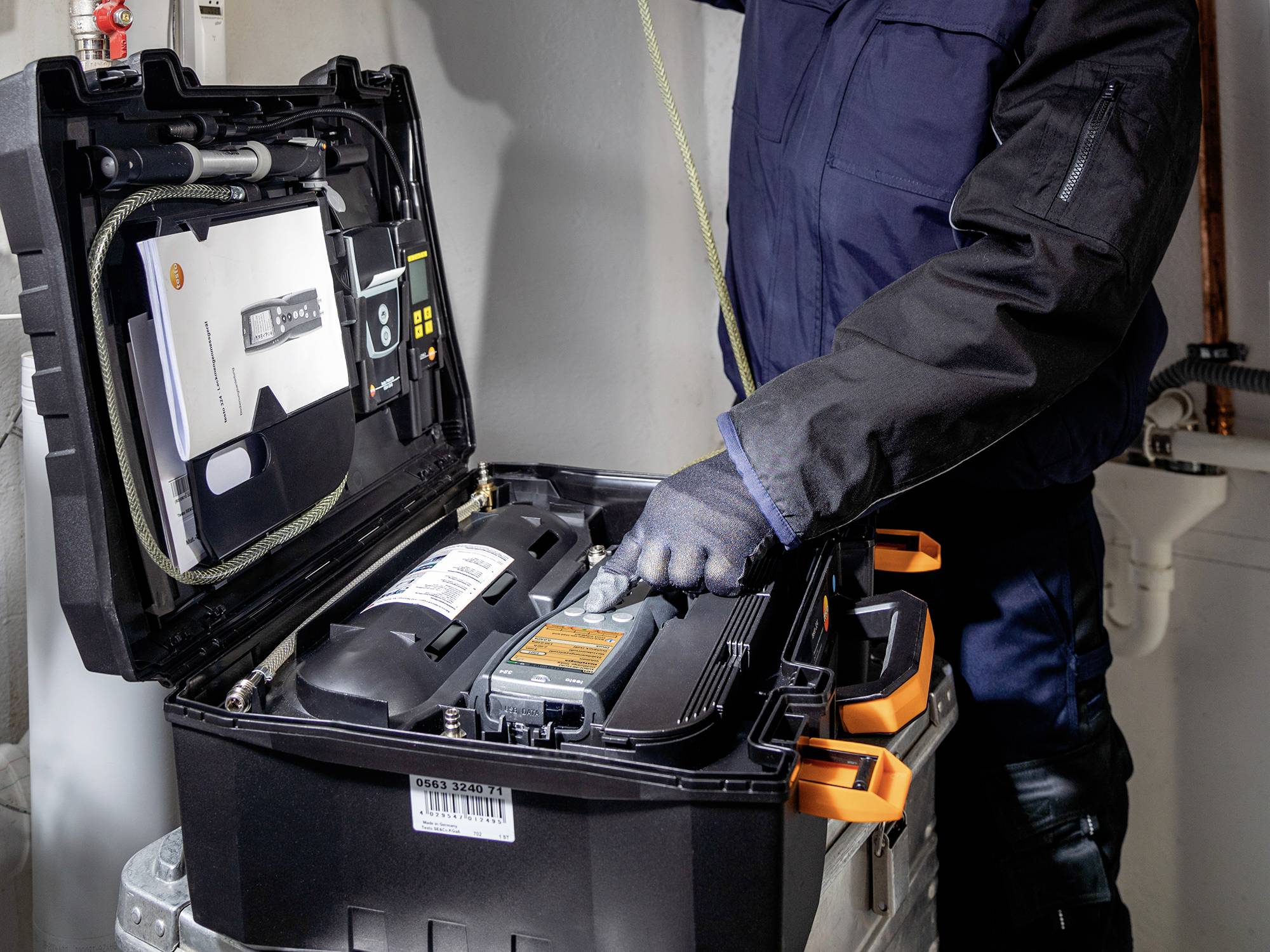 Techniker in blauer Arbeitskleidung und Handschuhen überprüft einen geöffneten Werkzeugkoffer mit Messtechnik in einem Haus.