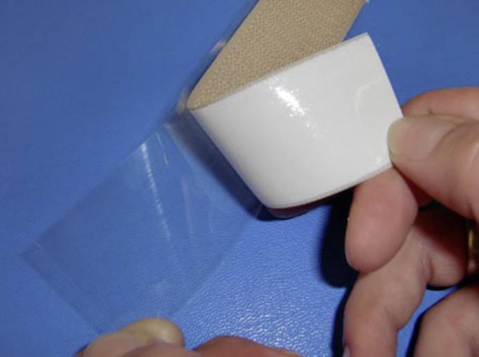 Zwei Hände halten ein Klettband über einer blauen Arbeitsfläche und lösen die Schutzfolie der rückseitigen Klebefläche.