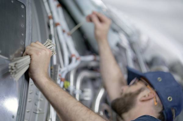 Ein Techniker installiert Kabelbinder in einem Flugzeug, um Kabel zu organisieren. Er trägt blaue Arbeitskleidung und Schutzbrille.