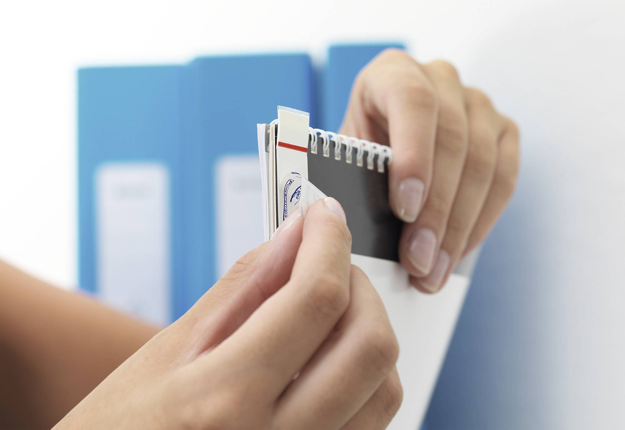 'Nahaufnahme von Händen, die ein Farbmuster-Buch vor blauen Aktenordnern durchblättern. Auswahl von Farben oder Materialien.'