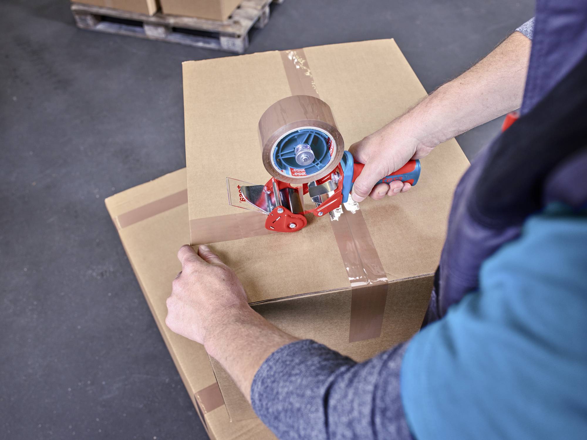 Eine Person klebt mit einem Abroller Klebeband auf einen Karton. Im Hintergrund steht ein weiterer Karton auf einer Palette.