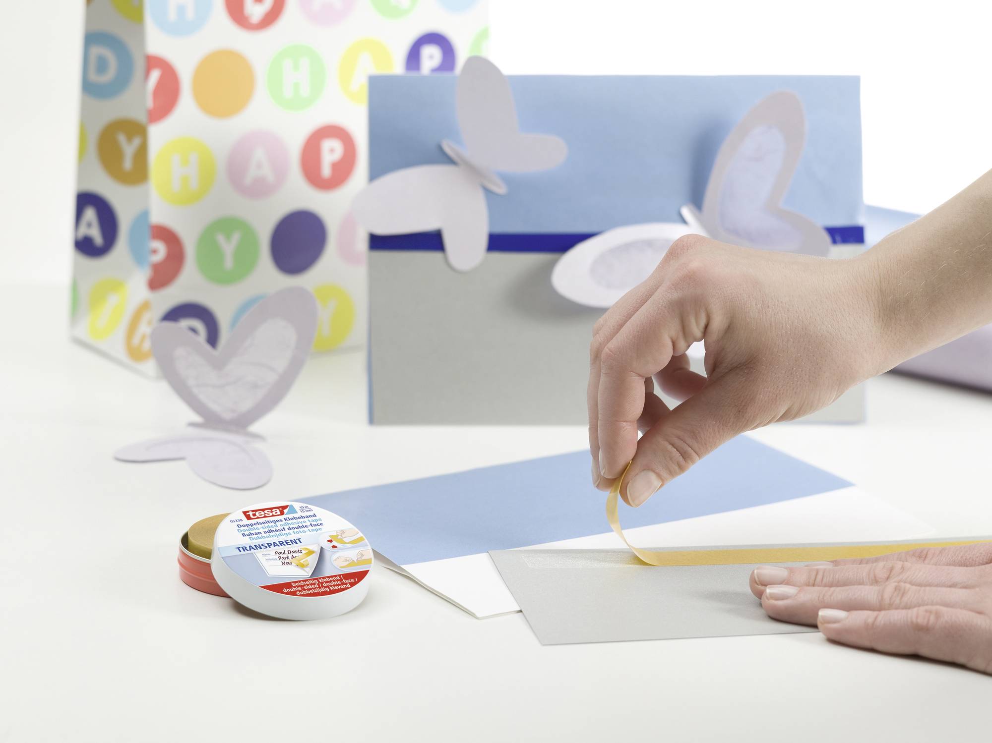 Eine Hand klebt mit einem doppelseitigen Klebeband Schmetterlingsdekorationen auf eine Karte. Im Hintergrund eine Geschenktüte.