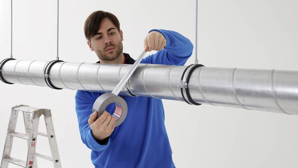 Ein Mann in blauem Pullover steht auf einer Leiter und klebt einen langen grauen Metallrohr mit Silbertape ab.