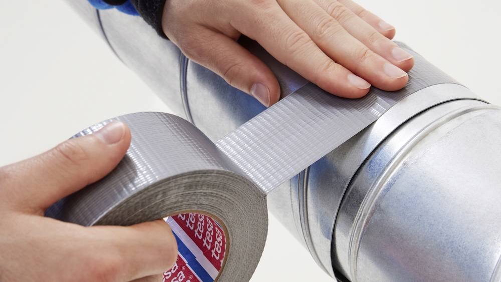 Jemand repariert ein rundes Metallrohr mit silbernem Klebeband, indem er es um eine Verbindung wickelt.